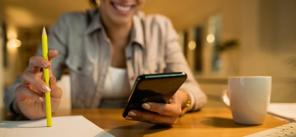 Mulher sorridente segura um lápis amarelo e usa o celular em uma mesa com café, focada em seu planejamento pessoal.