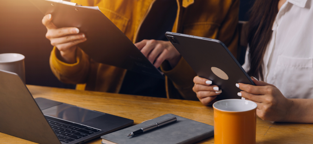 Duas pessoas trabalham em uma mesa de madeira usando tablet, prancheta e notebook, ao lado de canecas e uma agenda.