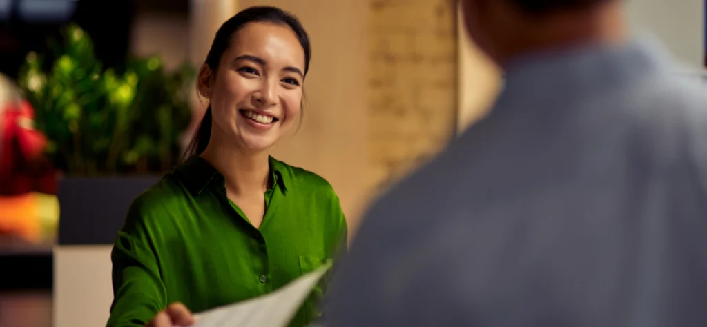 Mulher asiática sorridente de camisa verde entrega um documento a um colega desfocado, em um ambiente corporativo.