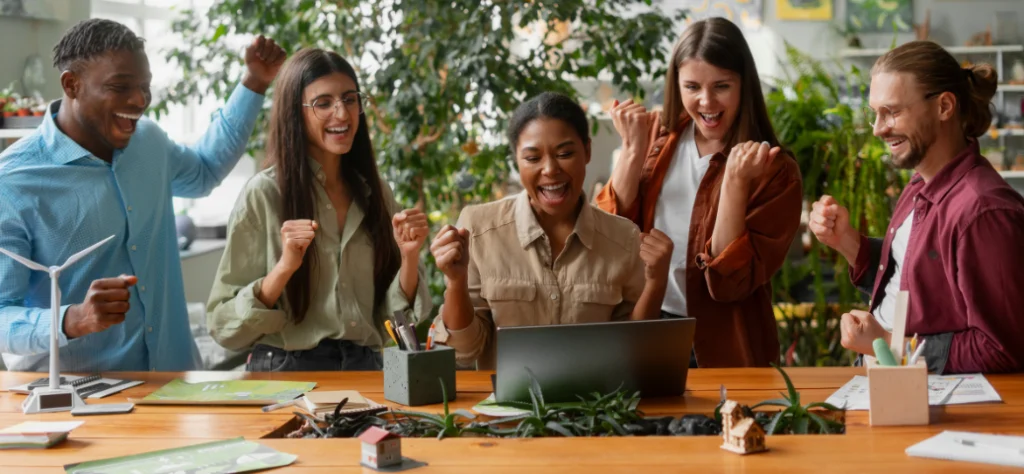 Equipe de trabalho diversa comemora com entusiasmo em frente a um notebook, demonstrando sucesso, união e colaboração no escritório.
