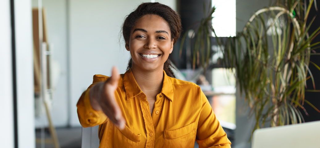 Mulher negra sorridente estende a mão para um aperto de mão em um escritório, transmitindo boas-vindas e profissionalismo.