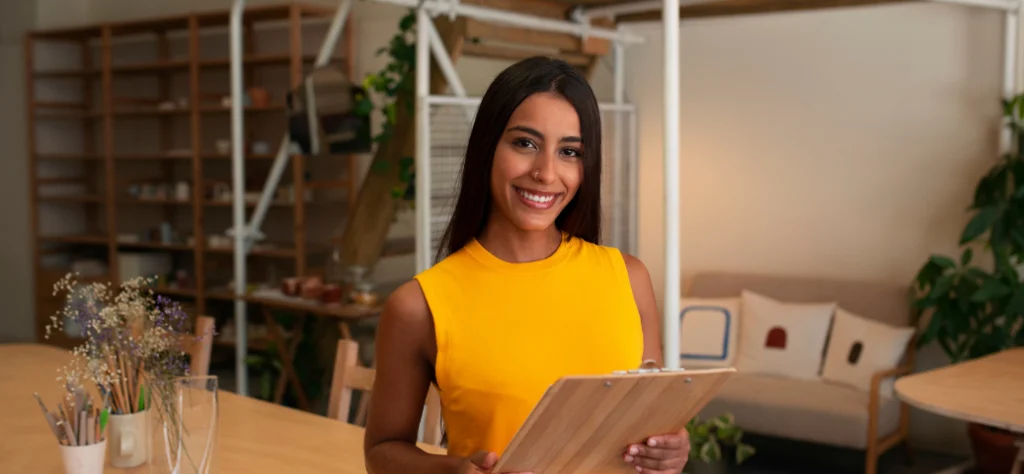 Jovem mulher sorridente com regata amarela segura uma prancheta de madeira em um escritório moderno, arborizado e bem iluminado.