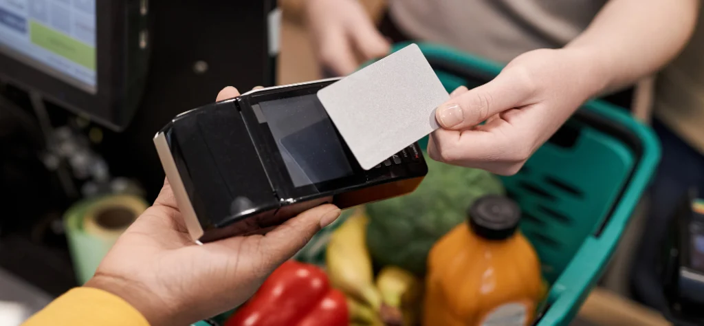 Close-up de uma mão aproximando um cartão cinza de uma máquina de pagamento sobre um cesto com frutas e legumes.
