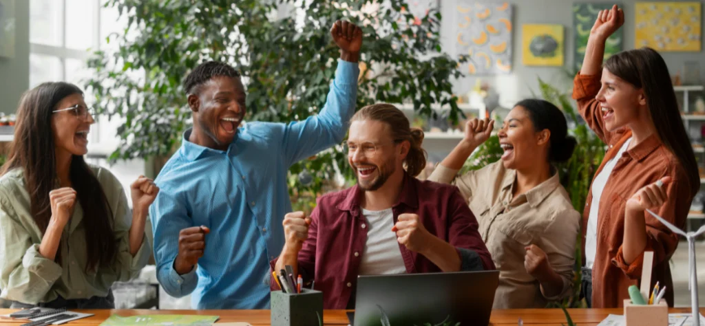 Equipe diversa comemora alegre ao redor de mesa com notebook em escritório, vibrando com braços levantados e punhos cerrados.
