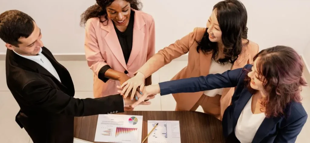 Quatro colegas de trabalho, um homens e três mulheres, estão empilhando as mãos uns sobre os outros em uma mesa. Eles estão sorrindo e usando blazers. Há papéis e um lápis na mesa.
