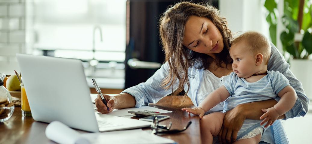 Mulher trabalha em notebook e escreve enquanto segura um bebê no colo, em uma mesa com calculadora e documentos.