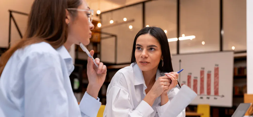 Duas mulheres em reunião em um escritório moderno; uma delas segura uma caneta e olha atentamente para a colega.