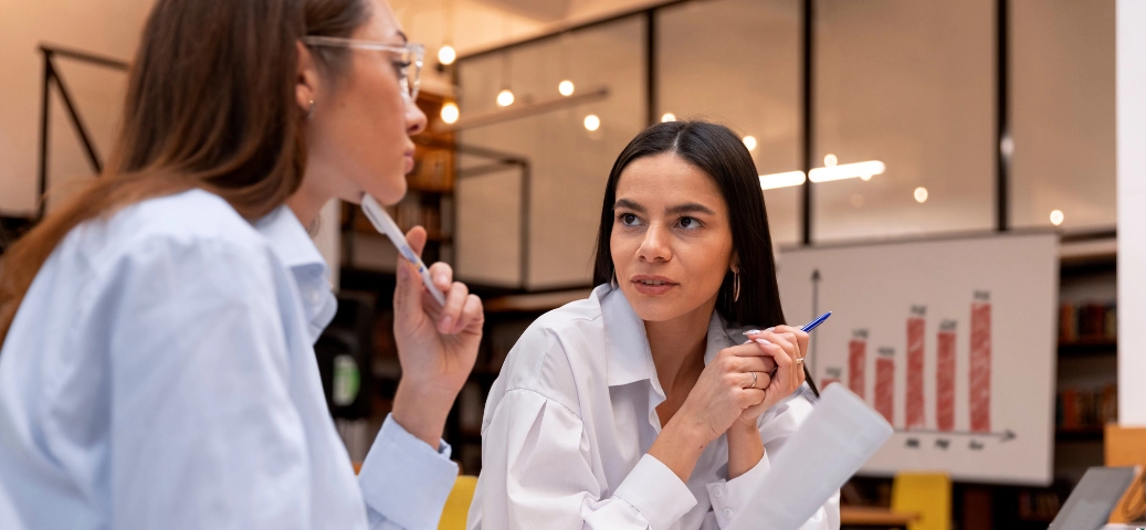 Duas mulheres em reunião em um escritório moderno; uma delas segura uma caneta e olha atentamente para a colega.