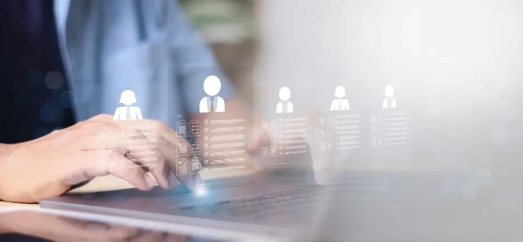 A imagem mostra as mãos de uma pessoa digitando em um laptop, com um foco suave na tela e nos dedos. No canto superior esquerdo da imagem, um vulto borrado sugere a presença de outra pessoa ou um fundo de escritório.