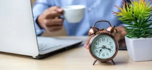 A imagem mostra uma cena em um escritório ou home office, com o foco principal em um despertador analógico de cor cobre, vintage, posicionado à direita no primeiro plano. O despertador tem um mostrador branco com números arábicos pretos e ponteiros pretos, e está obre uma mesa de madeira clara.