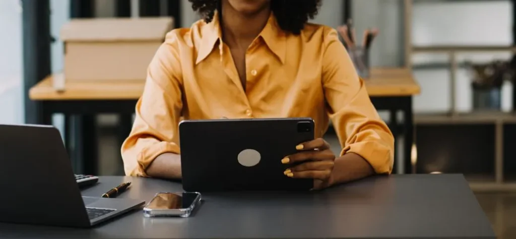 A imagem mostra uma pessoa sentada em uma mesa escura, provavelmente em um ambiente de escritório ou home office. O foco está na parte superior do corpo da pessoa, que veste uma camisa de manga comprida de cor mostarda ou dourada com um colarinho proeminente. A pessoa tem cabelos curtos e encaracolados, e unhas pintadas de amarelo.