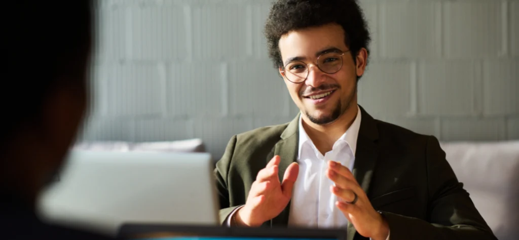 Homem de óculos e blazer verde gesticula e sorri durante reunião, com notebook e outra pessoa em primeiro plano.