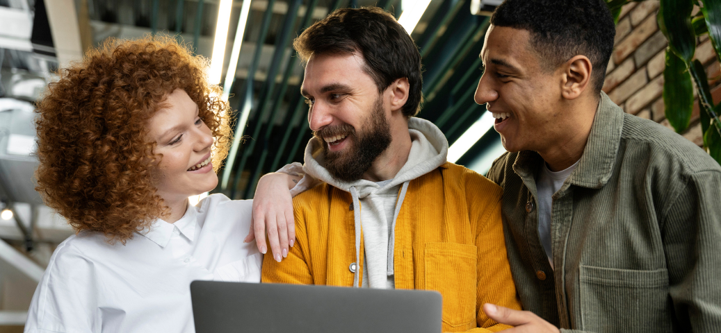 Três colegas de trabalho sorriem enquanto olham para um notebook, demonstrando integração e colaboração em equipe.