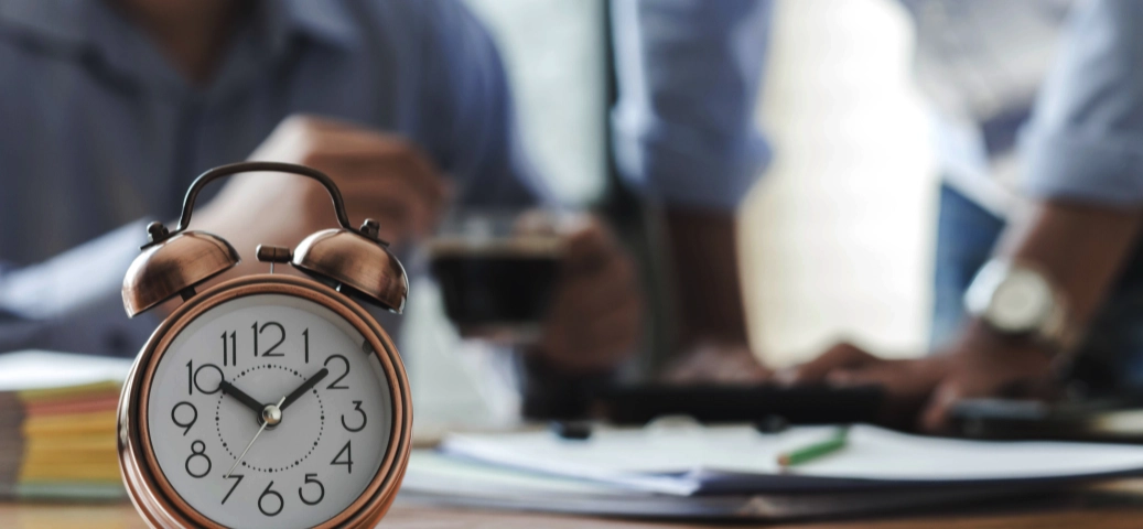 Despertador vintage em foco sobre mesa de escritório, com pessoas e papéis desfocados ao fundo. Gestão de tempo.