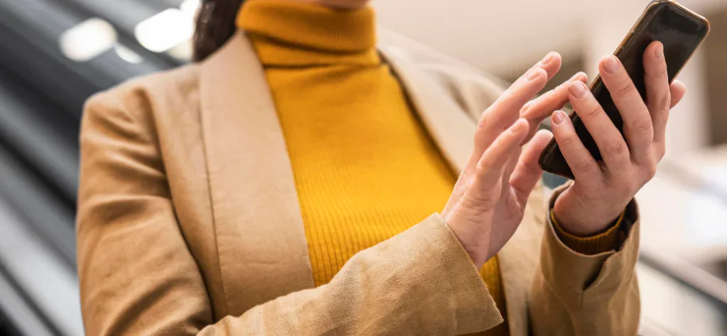 Close-up das mãos de uma pessoa usando blazer bege e blusa de gola alta amarela operando um smartphone.