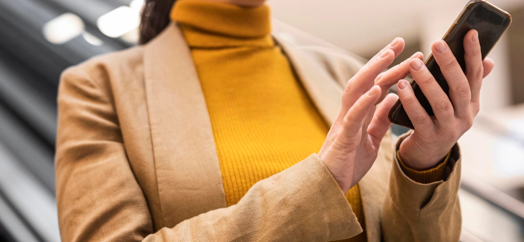 Close-up das mãos de uma pessoa usando blazer bege e blusa de gola alta amarela operando um smartphone.