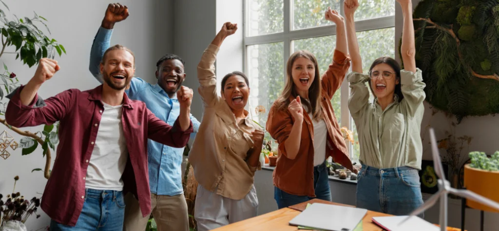 Equipe diversa comemora alegre com os braços levantados em um escritório moderno, expressando sucesso e vitória.