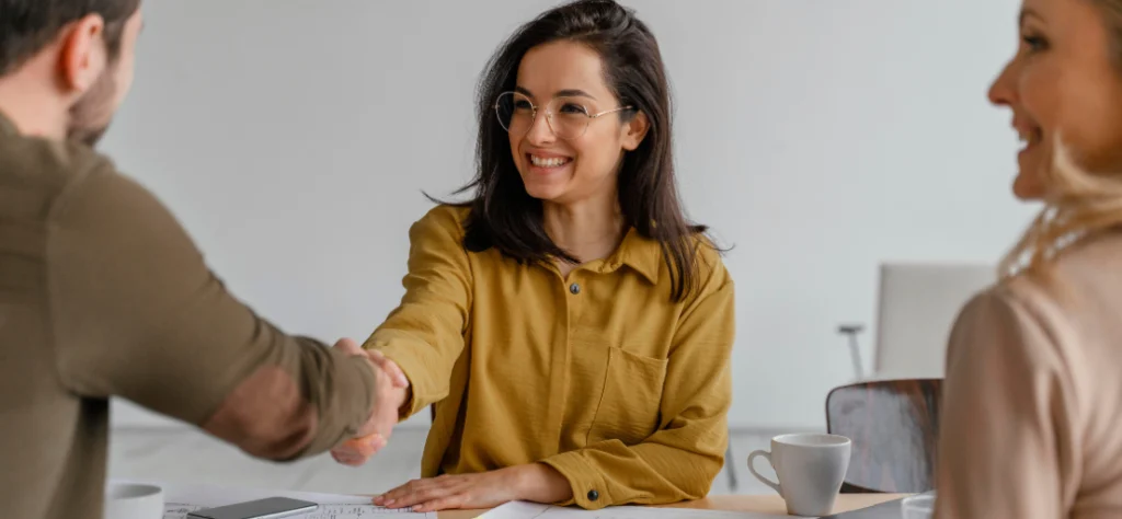 Mulher de óculos e camisa amarela sorri ao apertar a mão de um colega em uma reunião, simbolizando parceria e novos acordos.