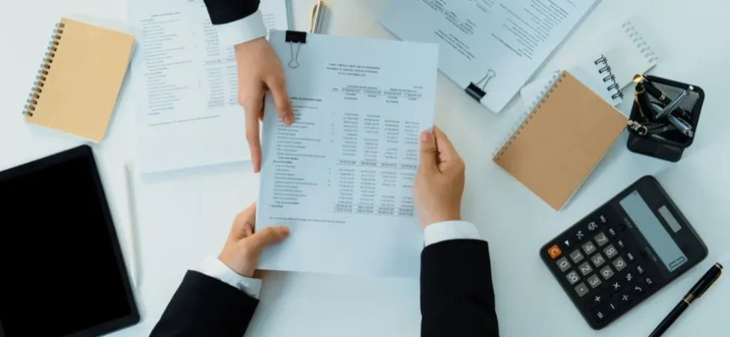 Duas mãos em trajes profissionais trocam um documento financeiro sobre uma mesa desorganizada com uma calculadora, um tablet e blocos de notas.