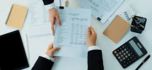 Duas mãos em trajes profissionais trocam um documento financeiro sobre uma mesa desorganizada com uma calculadora, um tablet e blocos de notas.
