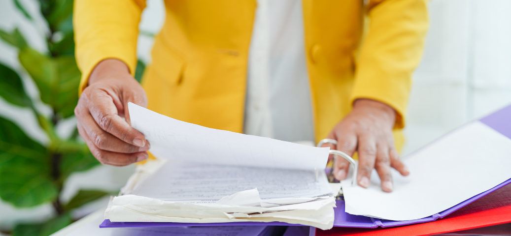 Pessoa de blazer amarelo organiza papéis dentro de uma pasta fichário aberta, demonstrando gestão de documentos.