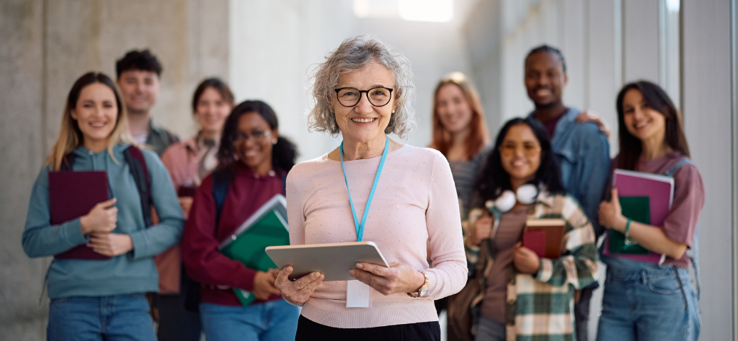 Professora veterana sorridente segurando um tablet à frente de um grupo diversificado de estudantes em um corredor.