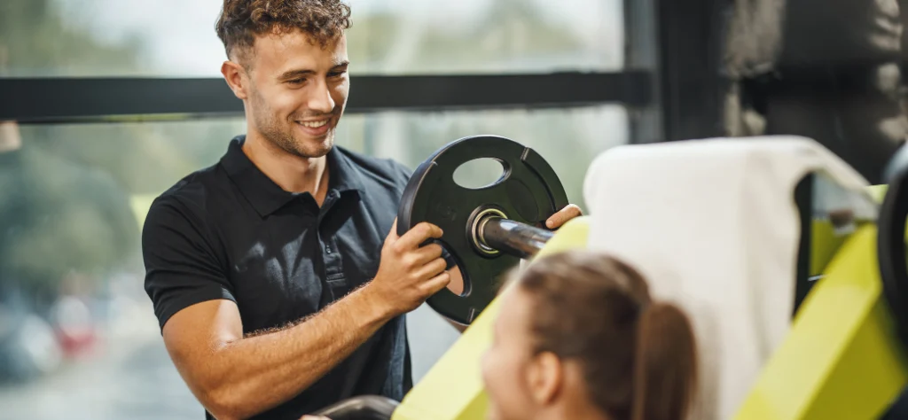Instrutor sorri ao colocar anilha de peso em aparelho de musculação para aluna treinar na academia.