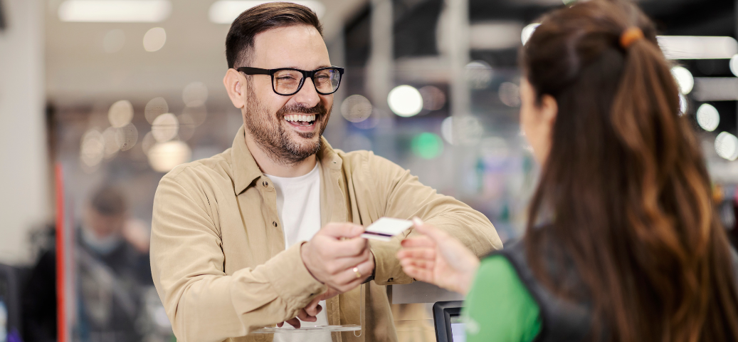 Homem sorridente de óculos entrega cartão de crédito para atendente em um ponto de venda comercial.