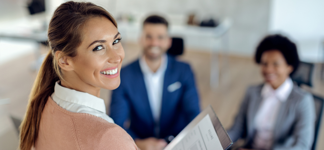Mulher sorridente segura currículo em primeiro plano, com dois recrutadores desfocados ao fundo em uma sala de entrevista.