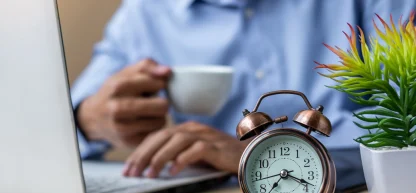 Desfoque de homem trabalhando em laptop, com um despertador antigo em primeiro plano. Conceito de horas extras.