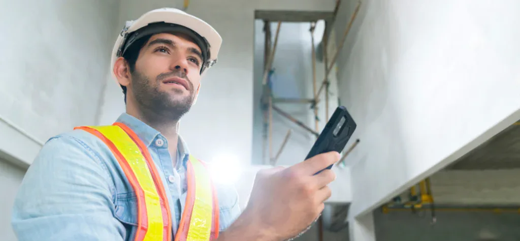 Trabalhador de capacete e colete reflexivo olha para cima enquanto segura um smartphone em uma obra em construção.