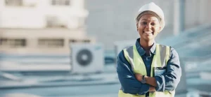 Ums trabalhadora confiante usando capacete e colete de segurança está de braços cruzados no telhado, com unidades de ar condicionado ao fundo.