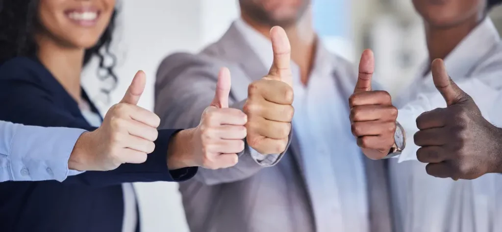 Um grupo diversificado de profissionais sorrindo e fazendo sinal de positivo, demonstrando trabalho em equipe e positividade em um ambiente de escritório iluminado.
