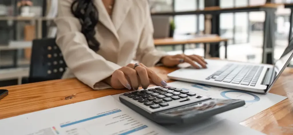 Uma mulher com um blazer claro opera uma calculadora enquanto trabalha em um laptop, com documentos financeiros em uma mesa de madeira em um escritório iluminado.