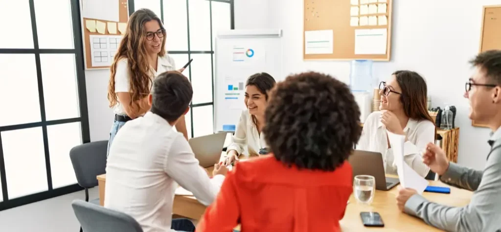 Um grupo diversificado de profissionais participa de uma reunião em uma mesa de conferência, discutindo documentos com um flip chart ao fundo.