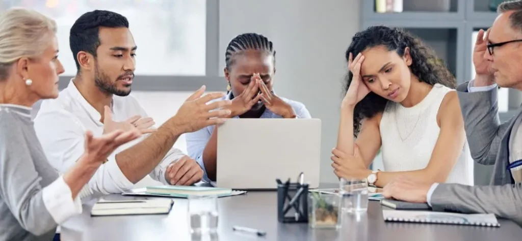 Foto de uma reunião de negócios tensa com cinco pessoas ao redor de uma mesa de conferência. Ao centro, uma mulher negra com tranças cobre o rosto com as mãos, aparentando estar angustiada ou sobrecarregada; um laptop está aberto à sua frente. À sua direita, uma mulher com longos cabelos escuros cacheados apoia a cabeça na mão, com uma expressão de cansaço e frustração. À esquerda da mulher angustiada, um homem de barba e camisa branca gesticula ativamente enquanto fala, parecendo estar em uma discussão acalorada ou tentando se explicar para uma mulher mais velha, de cabelos loiros curtos, que está à sua esquerda e também gesticula enfaticamente. No extremo direito da imagem, um homem mais velho de óculos e terno leva a mão à testa, com uma expressão de preocupação ou exasperação. Sobre a mesa, há cadernos, copos com água e material de escritório. A linguagem corporal de todos sugere um ambiente de conflito, estresse ou desacordo.