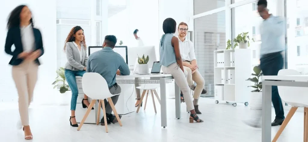 Um escritório moderno e iluminado com diversos profissionais colaborando em uma mesa, cercado por plantas e grandes janelas.
