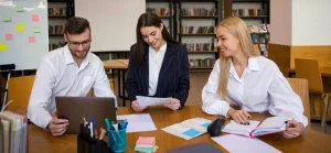 A imagem mostra três jovens, dois homens e uma mulher, sentados em uma mesa de madeira em um ambiente que parece ser uma biblioteca ou sala de estudos. À esquerda, um homem com barba e óculos está olhando para a tela de um notebook. Ele veste uma camisa branca de manga comprida. No centro, uma mulher de cabelo escuro, vestindo um blazer azul listrado sobre uma blusa branca, está segurando e lendo um documento. Ela tem um sorriso sutil. À direita, outra mulher, loira e com uma camisa branca de manga comprida, está sorrindo enquanto olha para o homem ou a mulher no centro. Ela tem um caderno aberto na mesa à sua frente e a mão direita sobre ele. A mesa está cheia de materiais de escritório, como canetas em um pote azul, papéis, adesivos coloridos e um mouse. Ao fundo, à esquerda, há um quadro branco com notas adesivas coloridas. À direita, prateleiras cheias de livros podem ser vistas, reforçando a ideia de um ambiente acadêmico ou de estudo. A iluminação é clara e o ambiente parece organizado.