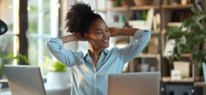 A imagem mostra uma mulher com a pele escura, sentada em uma mesa com dois notebooks, com os braços esticados para cima e as mãos atrás da cabeça, em uma postura de relaxamento ou alongamento. Ela tem os olhos fechados e um sorriso tranquilo no rosto, indicando um momento de alívio ou satisfação. A mulher usa uma camisa social clara, que parece ser azul-claro ou cinza-claro. Seu cabelo, de textura crespa, está preso em um coque alto e volumoso. O ambiente ao fundo sugere um escritório ou um espaço de trabalho em casa, com plantas em prateleiras e uma estante visível. A iluminação parece ser natural, vindo de uma janela, e atinge o rosto da mulher, criando um efeito de luz e sombra agradável. Há também uma luminária de mesa à esquerda, ligeiramente desfocada.