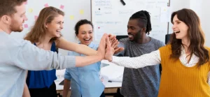 A imagem mostra cinco pessoas, três mulheres e dois homens, em um ambiente de escritório ou sala de reunião, participando de um "high five" coletivo, onde suas mãos se encontram no centro em um gesto de celebração ou trabalho em equipe. Todos estão sorrindo e parecem felizes ou entusiasmados.