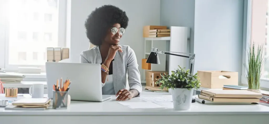Uma pessoa com um blazer cinza claro está sentada em uma mesa moderna com um laptop, plantas e materiais de escritório, imersa em pensamentos.