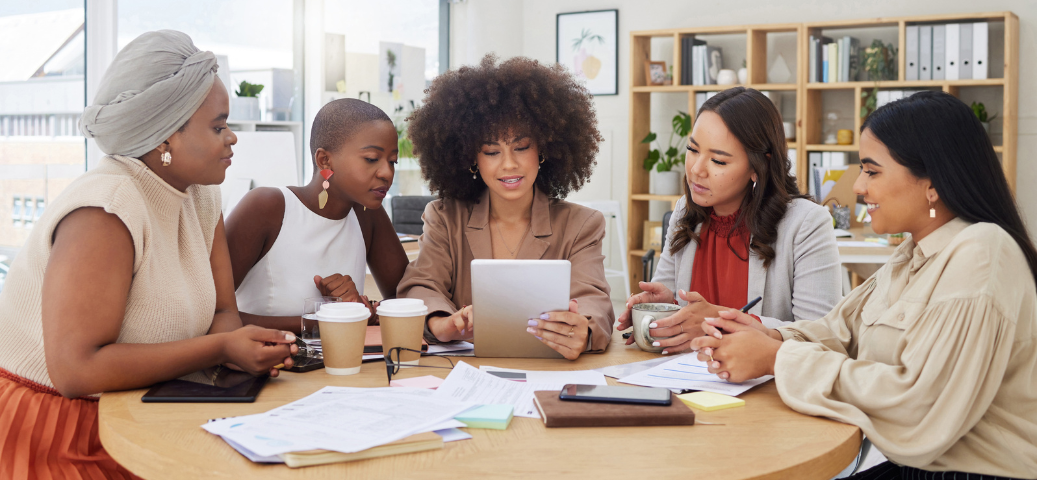 Aprendizado contínuo Um grupo de cinco mulheres conversa ao redor de uma mesa, com xícaras de café e documentos, em um ambiente de escritório moderno e iluminado.