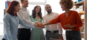 Uma equipe diversa de cinco profissionais, incluindo homens e mulheres, com diferentes etnias e tipos de cabelo, está reunida em um escritório. Eles sorriem e empilham as mãos uma sobre a outra no centro, transmitindo a ideia de união e trabalho em equipe.