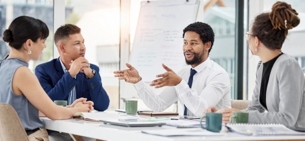 Um grupo diversificado de profissionais se envolve em uma discussão ao redor de uma mesa de conferência, com cadernos e xícaras de café em um ambiente de escritório moderno.