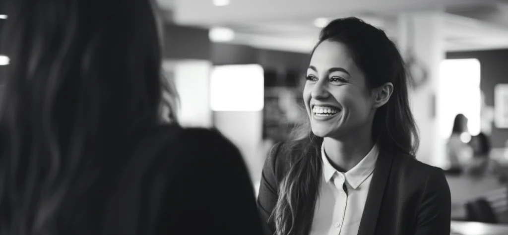 Uma mulher sorrindo, sentada à mesa e olhando para outra pessoa (parcialmente visível do ombro para trás). Ela está usando uma camisa social branca e um blazer escuro. A imagem está em preto e branco.