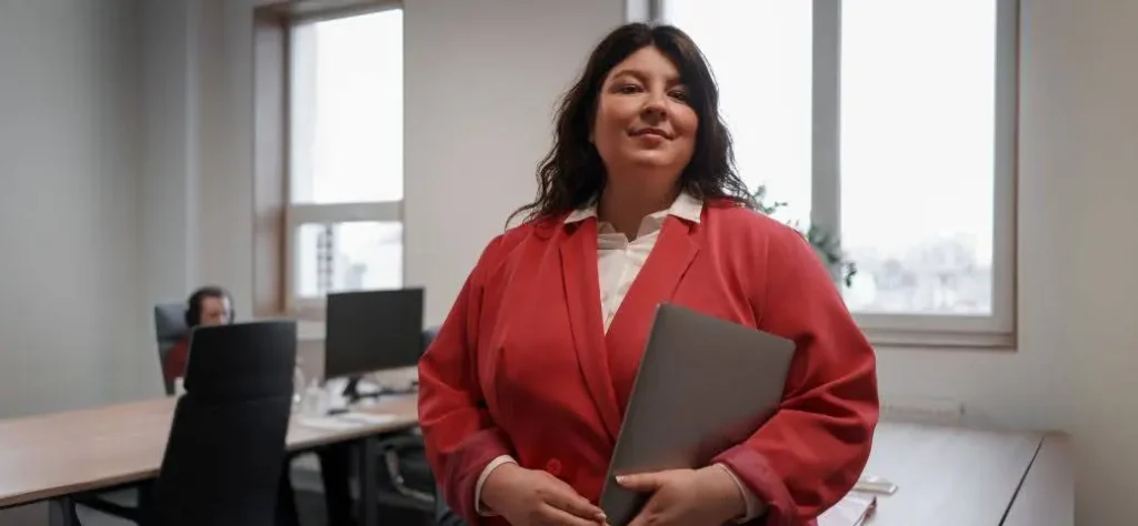 Uma pessoa usando um blazer vermelho está em pé com confiança em um escritório moderno com janelas grandes, segurando um laptop e olhando para a câmera.