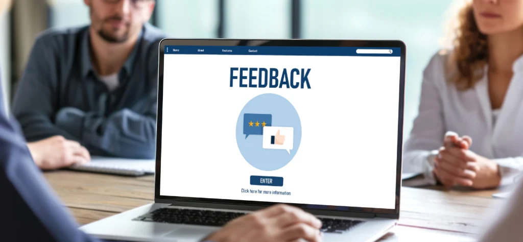 Uma visão aproximada de um laptop em uma mesa de conferência, com a palavra "FEEDBACK" exibida na tela. Abaixo da palavra, há dois balões de fala em um círculo azul claro, um com estrelas de avaliação e outro com um ícone de polegar para cima. O fundo mostra pessoas borradas sentadas ao redor da mesa.
