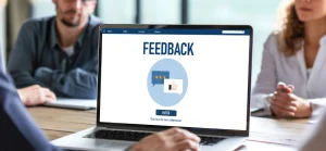 Uma visão aproximada de um laptop em uma mesa de conferência, com a palavra "FEEDBACK" exibida na tela. Abaixo da palavra, há dois balões de fala em um círculo azul claro, um com estrelas de avaliação e outro com um ícone de polegar para cima. O fundo mostra pessoas borradas sentadas ao redor da mesa.