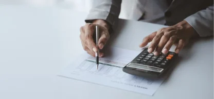 Duas mãos em trajes profissionais trocam um documento financeiro sobre uma mesa desorganizada com uma calculadora, um tablet e blocos de notas.