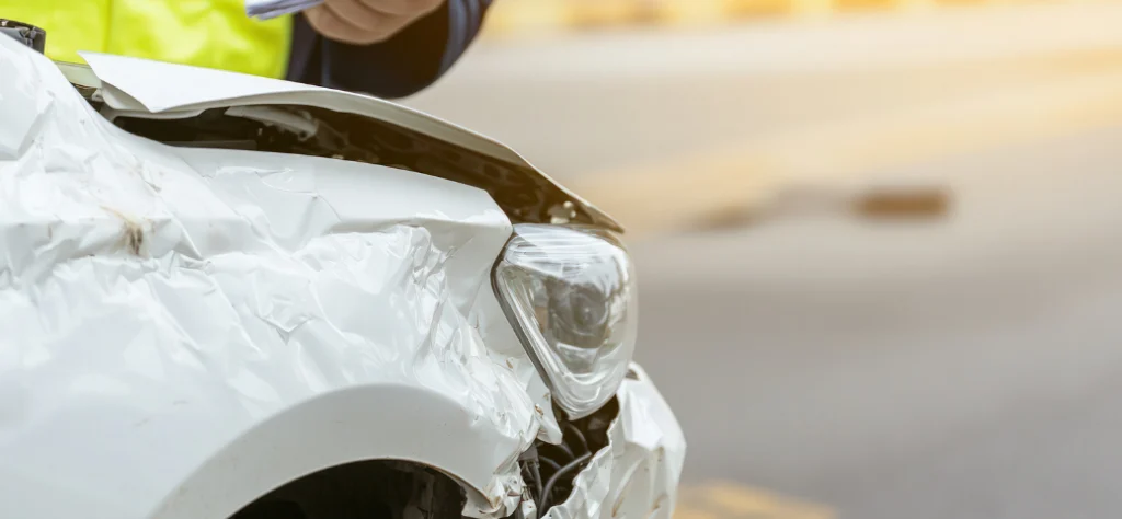 Close da parte frontal amassada de um carro branco após colisão na estrada, com pessoa de colete amarelo ao fundo.